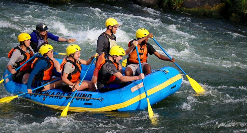 A group of students wearing safety gear paddle a raft through whitewater. 